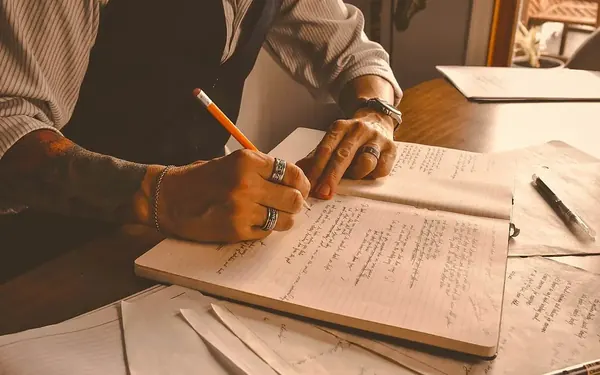 A writer drafting longhand in a notebook among loose pages
